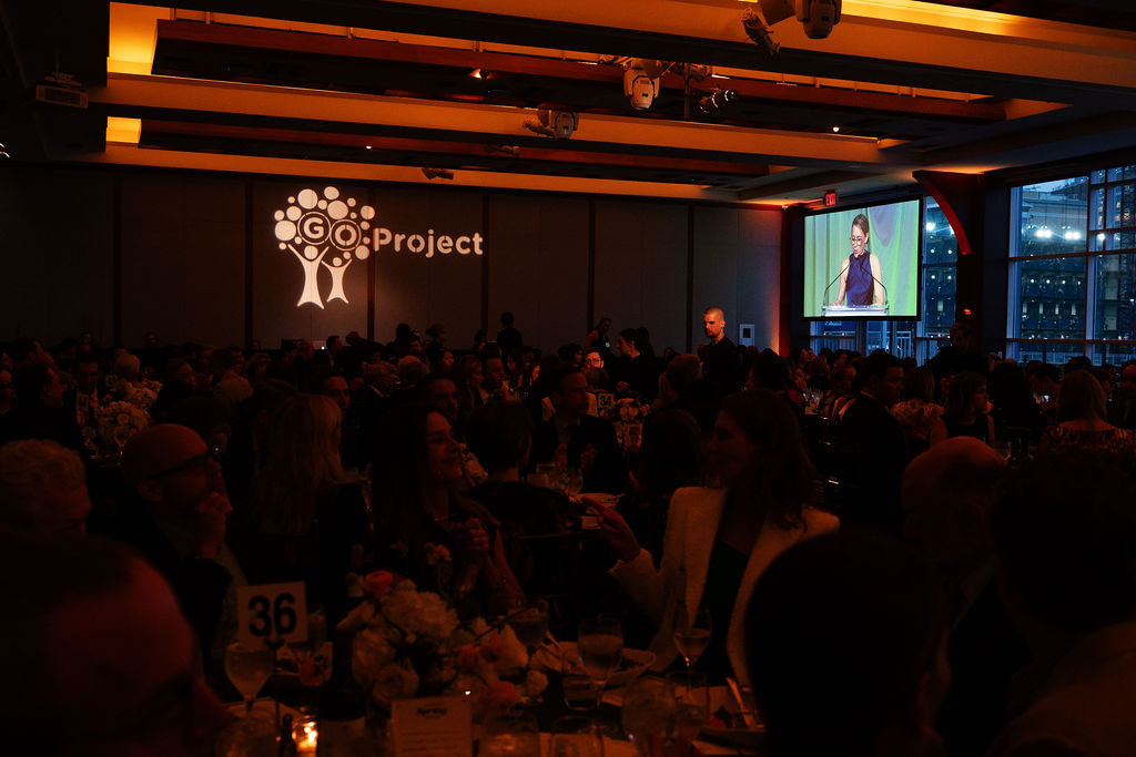 Party attendees sitting at round tables in a dim event hall look on as a speaker addresses them from the stage.
