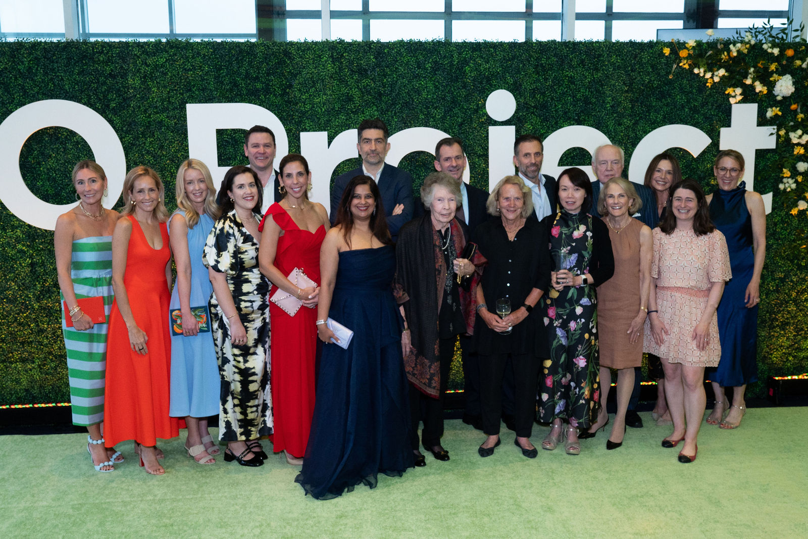 A large group of party attendees stand together in smile in front of an artificial hedge wall with large 'GO Project' lettering.