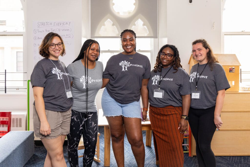 A group of women with grey shirts that say GO Project stand together and smile in a classroom.