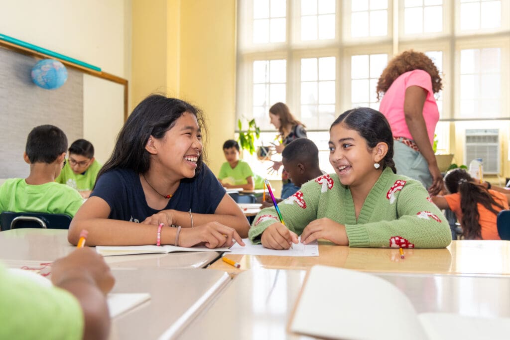 Two students at classroom desks smile at each other.