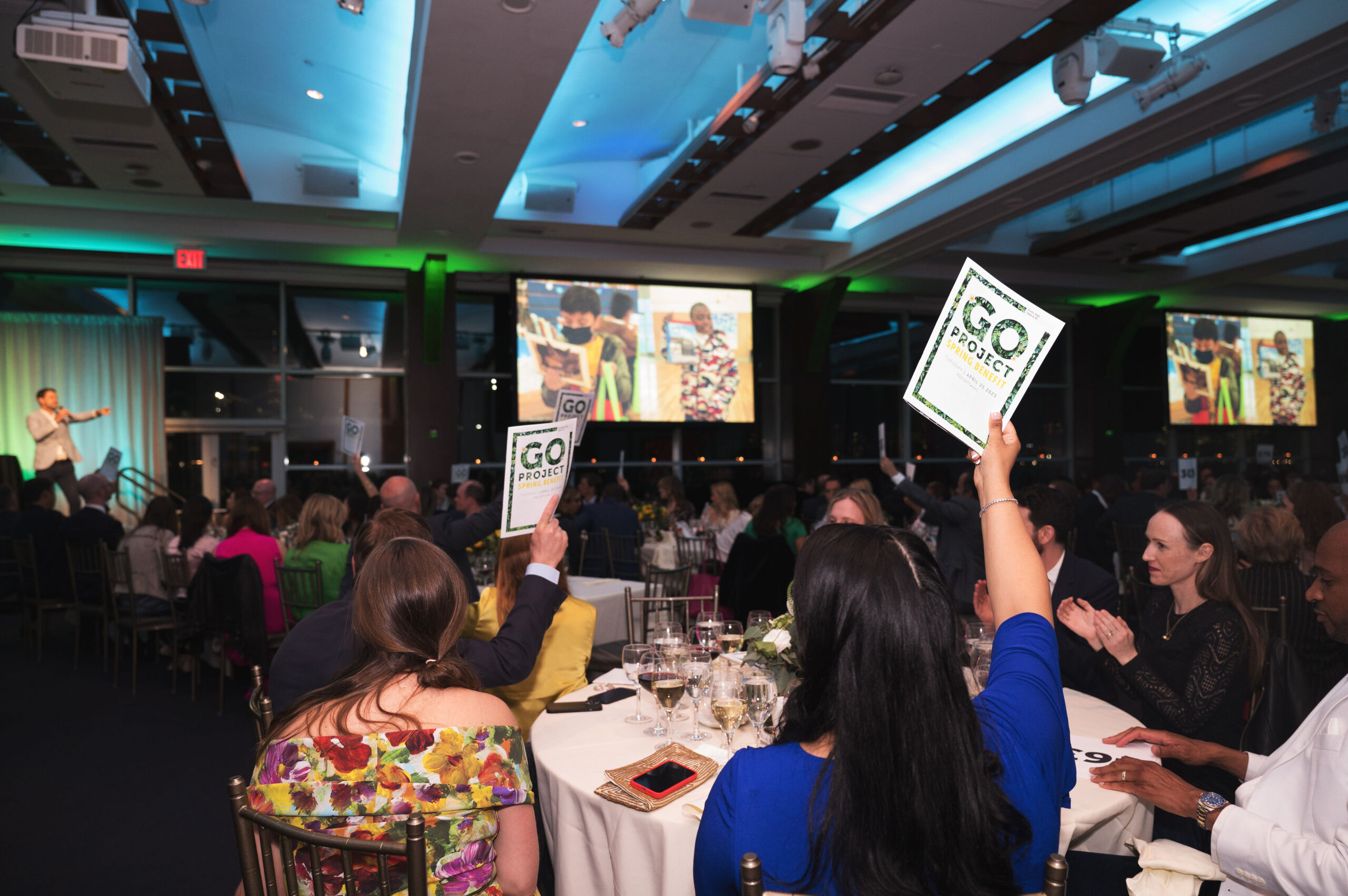 A dimly room full of people seated at round tabled. One woman, back to the camera, is holding up a booklet as if to bid in an auction.