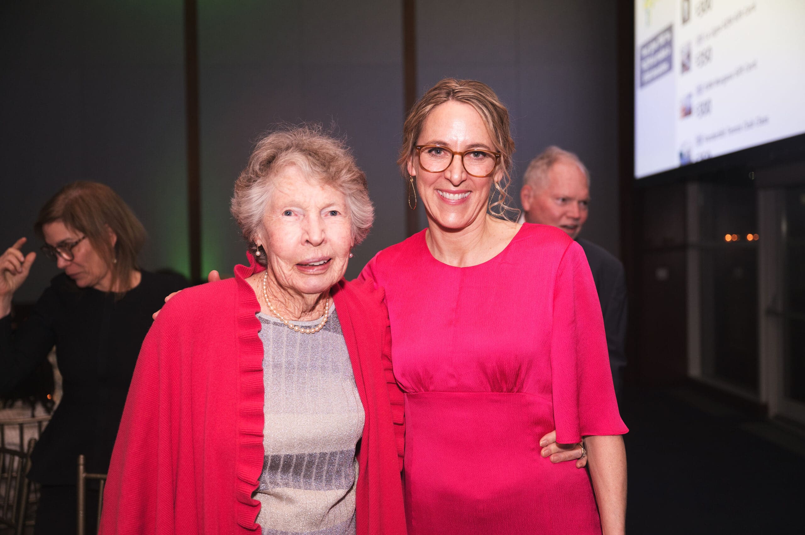 Two women stand together and smile with their arms around each other.