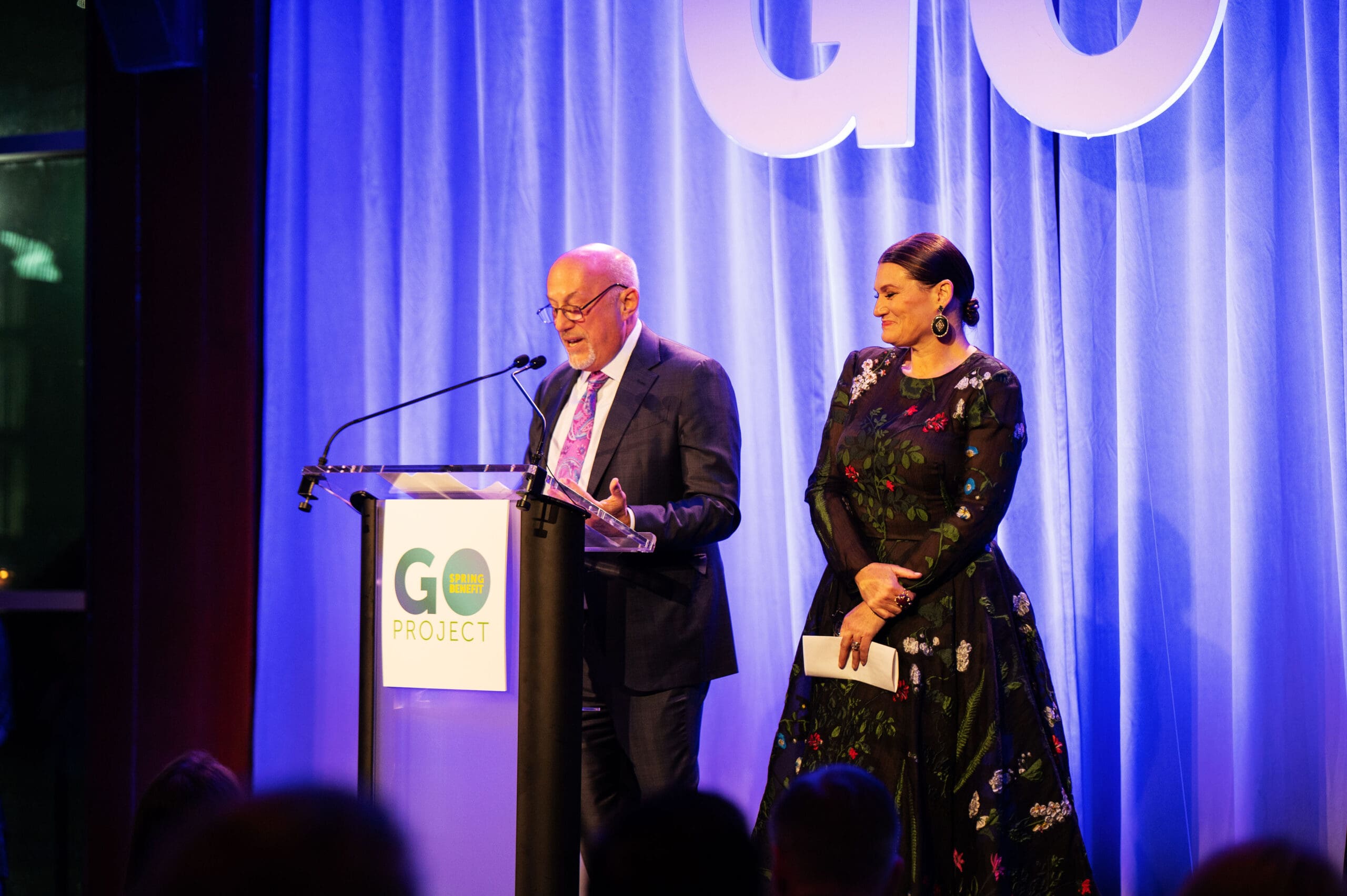 A man and a woman stand at a podium for GO Project's Spring Benefit.