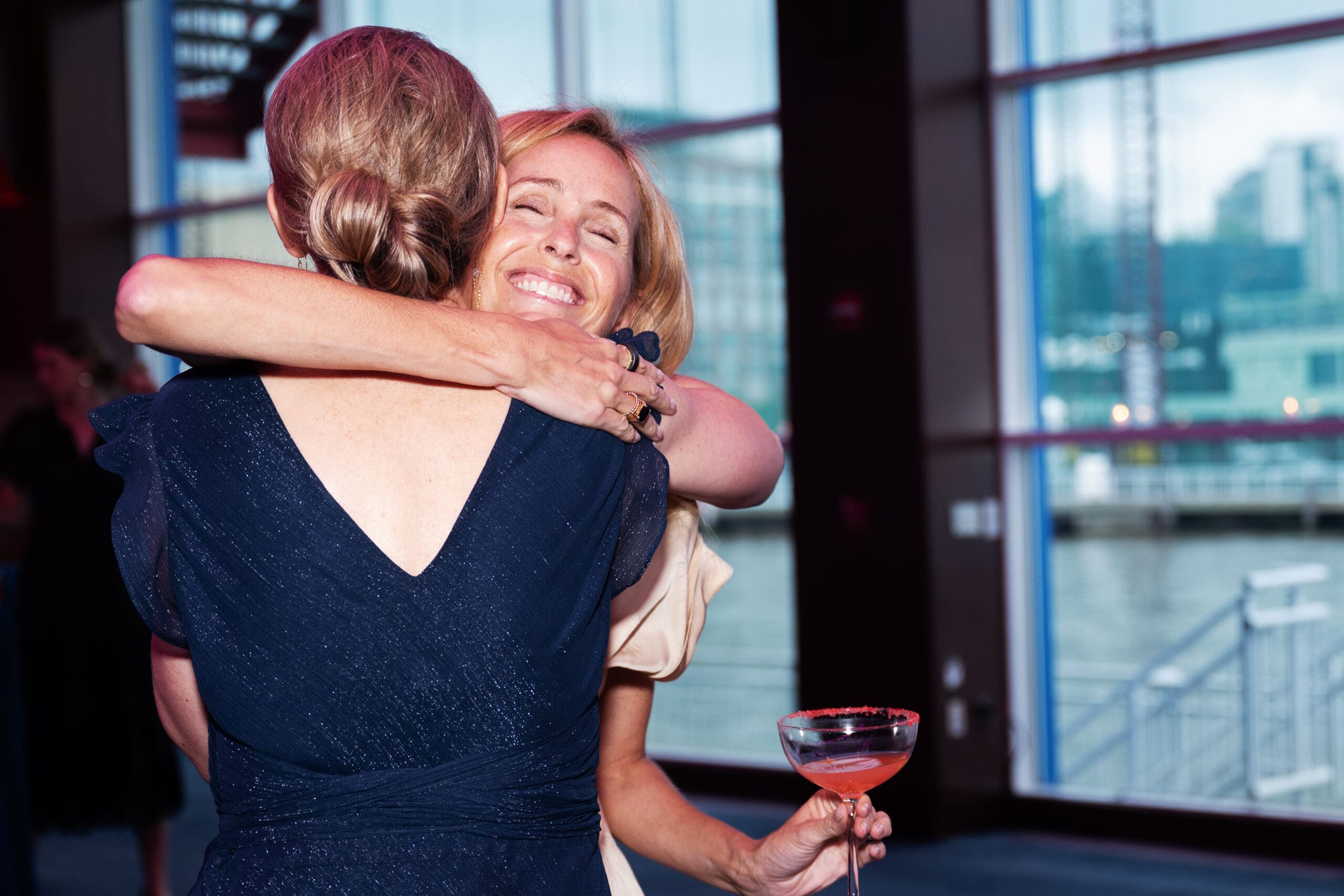 A woman with a pink-colored drink in her hand hugs another woman.
