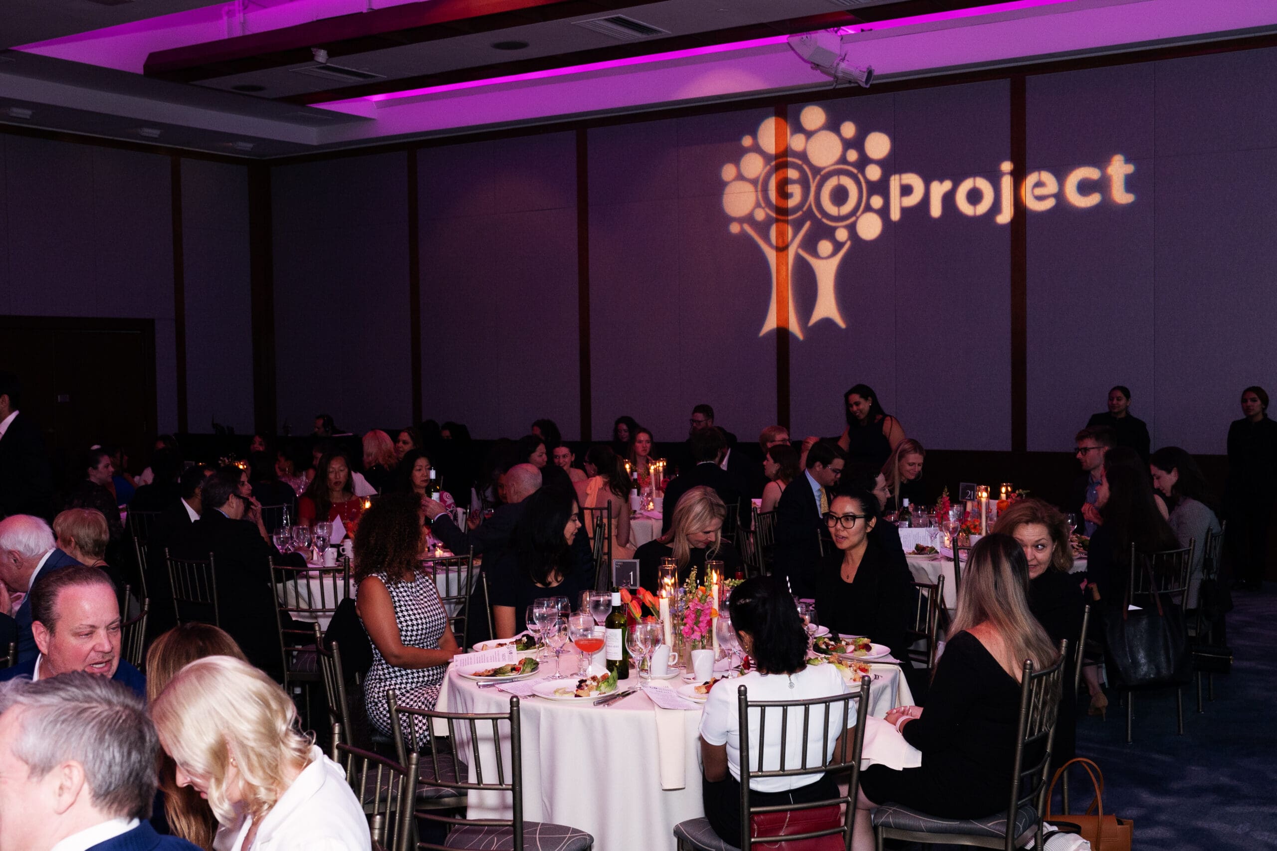 Party attendees at round tables in a dim event hall converse with one another.