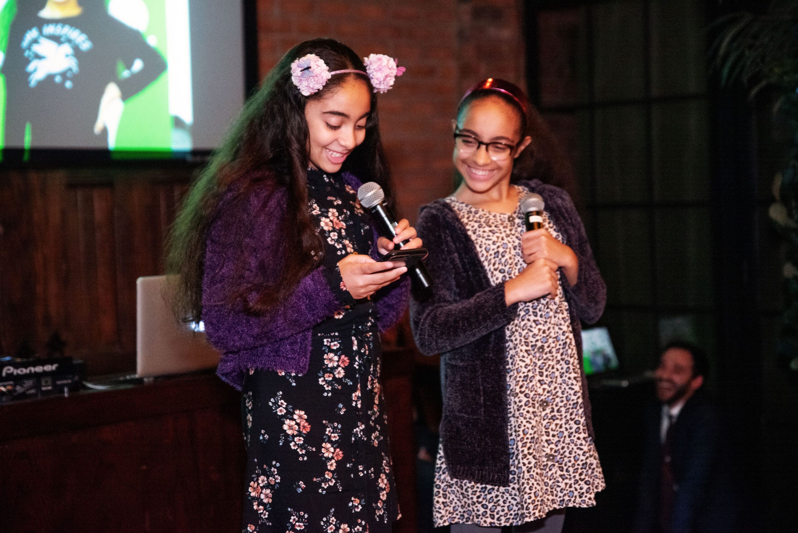 Two young girls holding microphones smile and address the room.