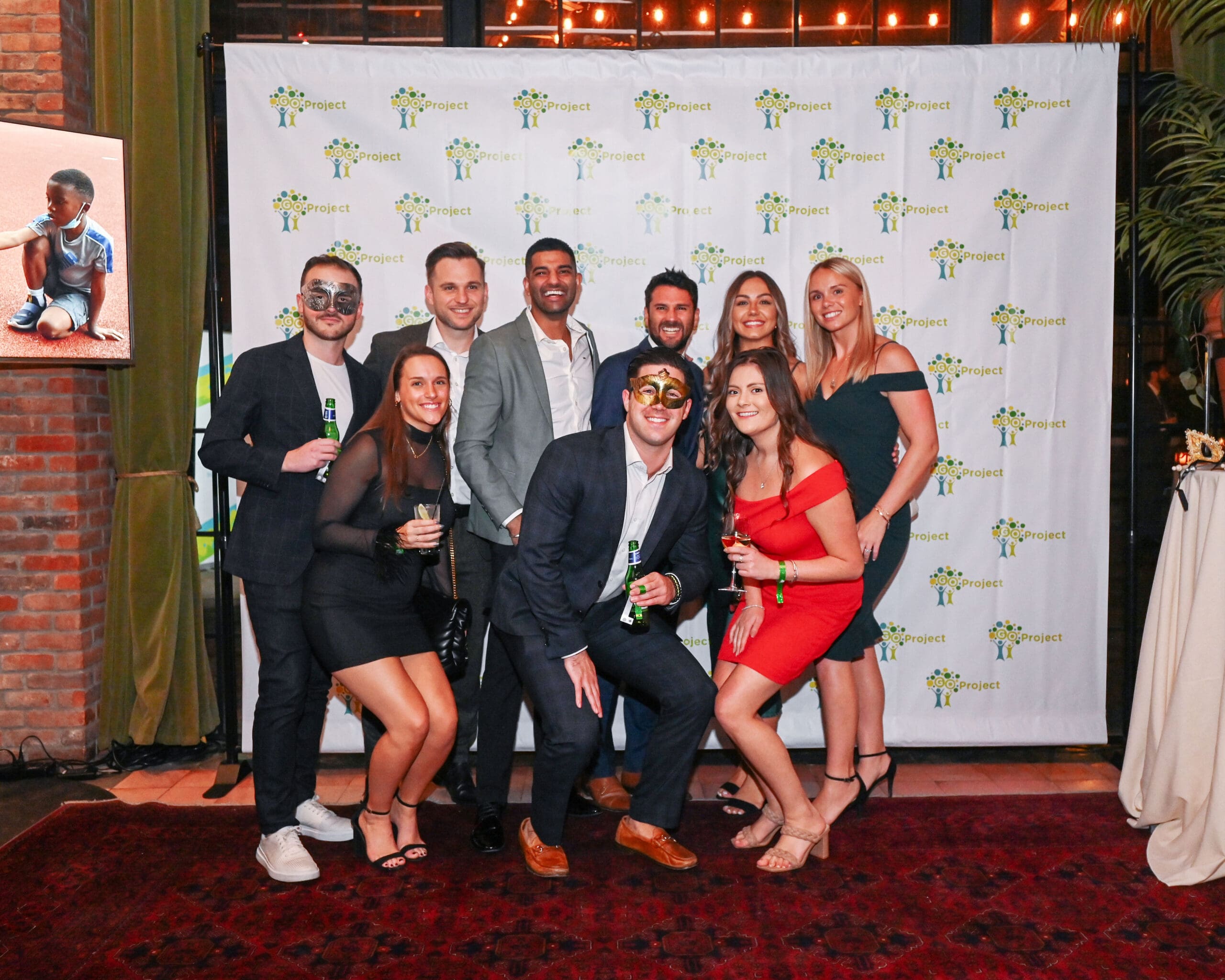 A group of attendees in cocktail attire, some with masquerade masks, stand together in front of a GO Project photo wall.