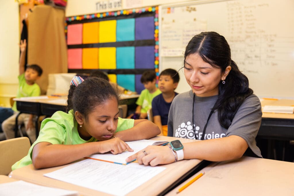 An instructor and a student work on classwork together at a desk in a classroom.