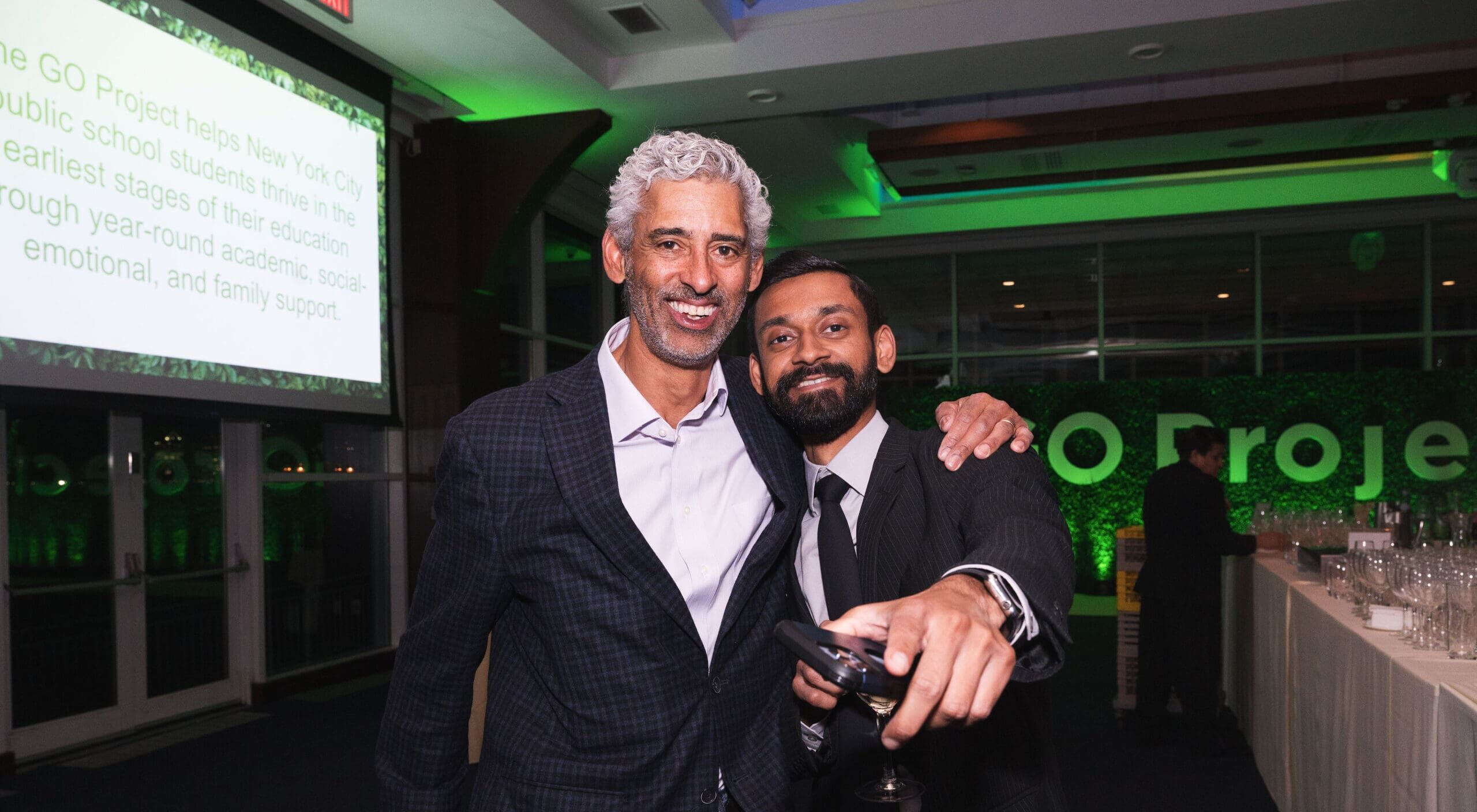 Two men stand together and smile, with one man pointing ahead of him.