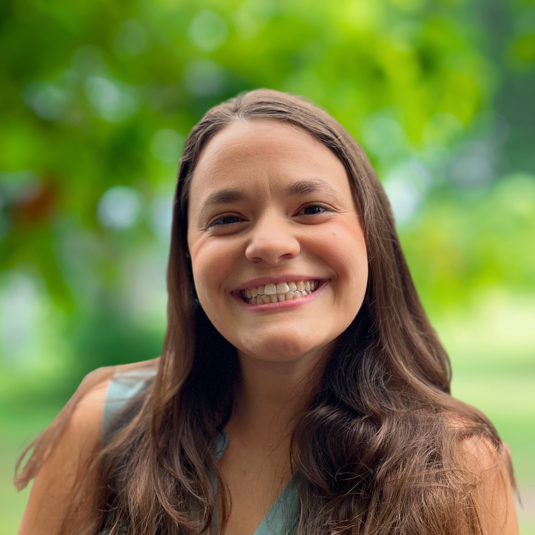 Sara Palombo smiles in front of a hazy tree-filled background.