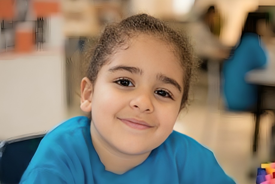Merna, who is wearing a blue shirt, smiles from her seat in a classroom.