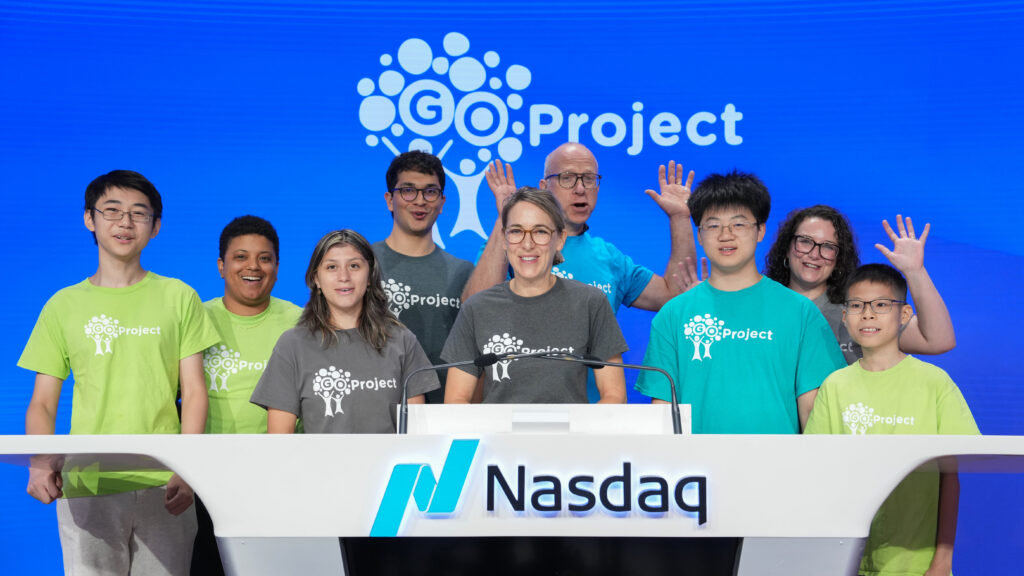 Adult and teenagers in bright green shirts are smiling and posed behind a Nasdaq podium.