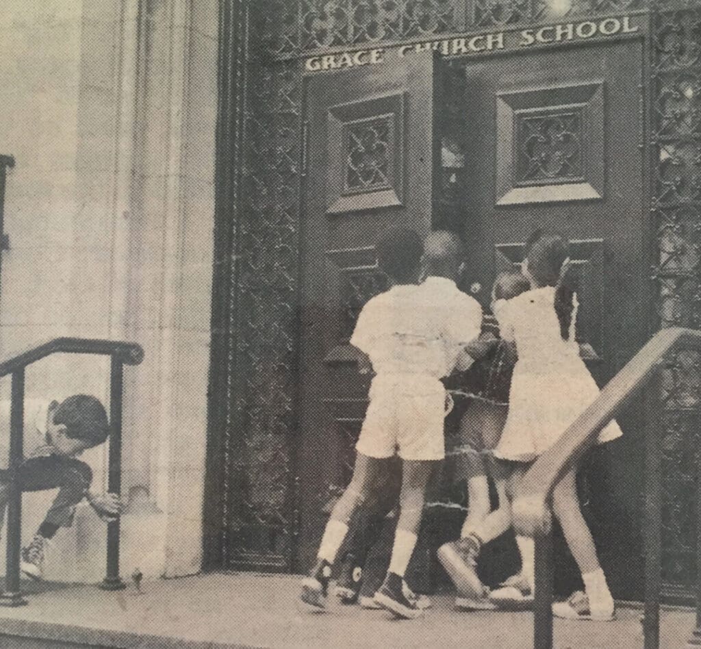A group of young students stand at the door of Grace Church School as it is opened by a teacher in a historical photograph.