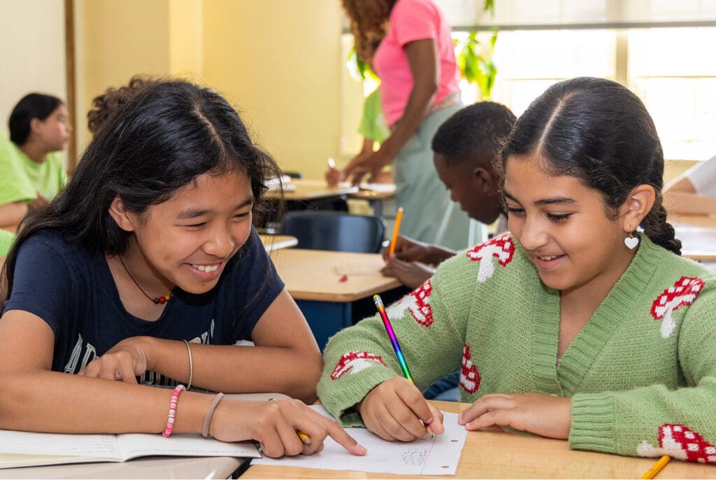 Two students at classroom desks smile and complete classwork together.