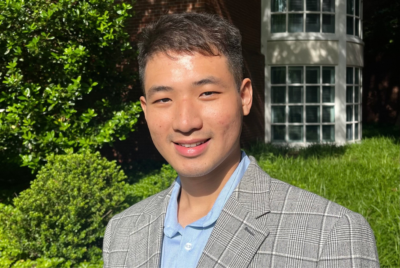 Zhen smiles in front of a brick-facade building and green foliage.